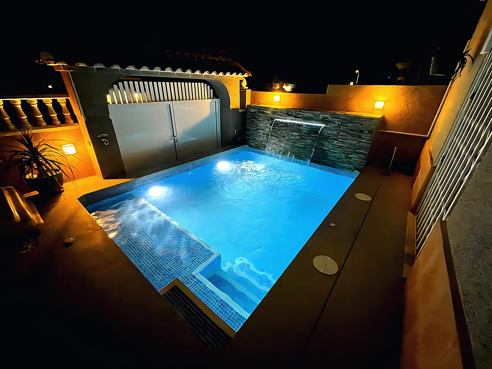 Square pool lit at night, pale blue mosaic, water blade on a slate-clad wall and warm lanterns, built by ORIGEN, Costa Blanca