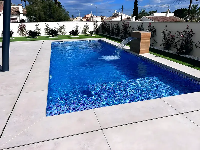 Rectangular pool with deep blue mosaic, timber-look waterfall column, light grey porcelain terrace and a planted white wall