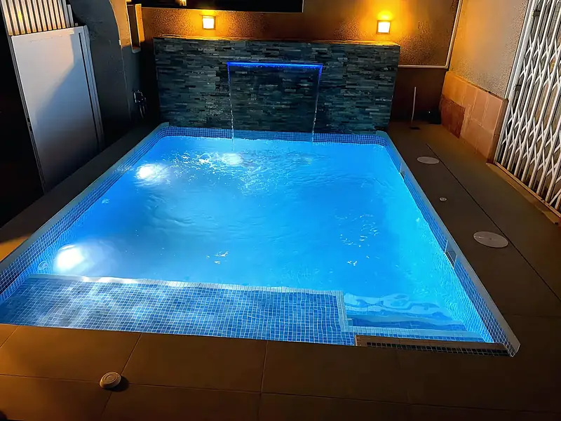 Square pool at night with a blue LED water blade on a slate feature wall, pale blue mosaic and warm wall lights