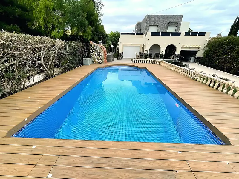 Pool with a rounded end and blue mosaic, composite wood decking all round, stone balustrade and a villa with arches behind