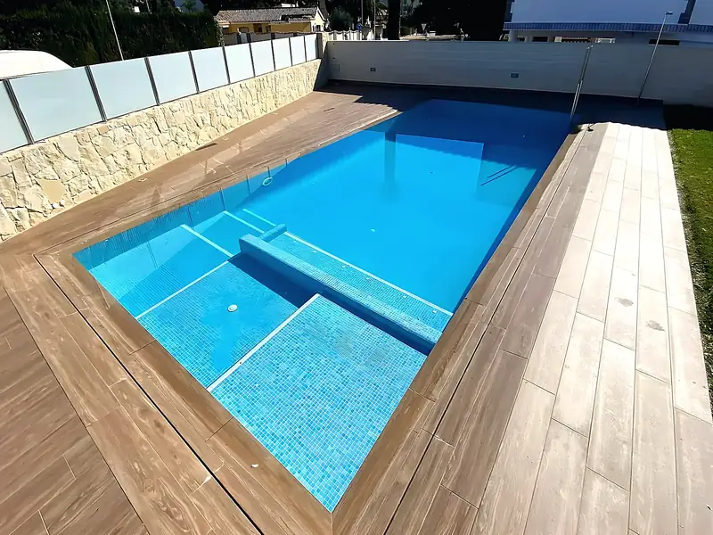 Pool with light blue mosaic, sun shelf and underwater bench, wood-effect porcelain deck, stone wall and frosted glass fence
