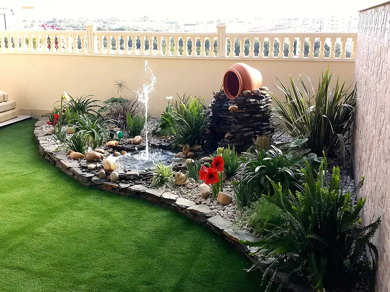 Garden with artificial lawn, slate-edged raised bed, small pond with fountain jet, terracotta urn, ferns and red amaryllis