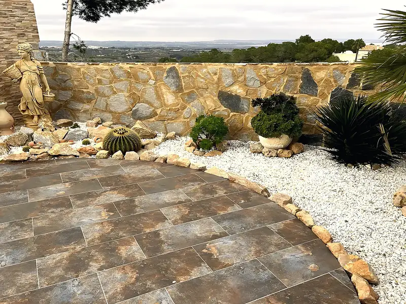 Curved terrace in rust-toned tiles edged with natural stone, white pebble beds with cacti, a statue and a natural stone wall