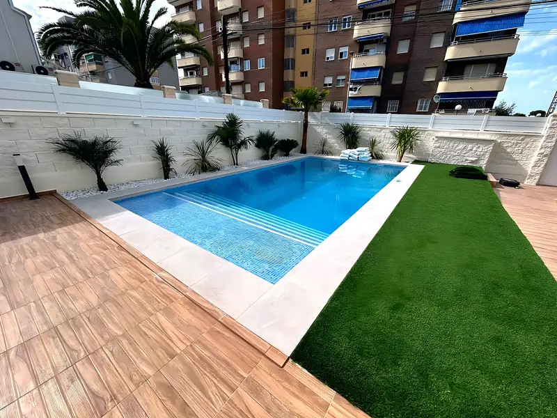 Pool with light blue mosaic and full-width steps, wood-effect porcelain deck, artificial lawn and palms on white pebbles