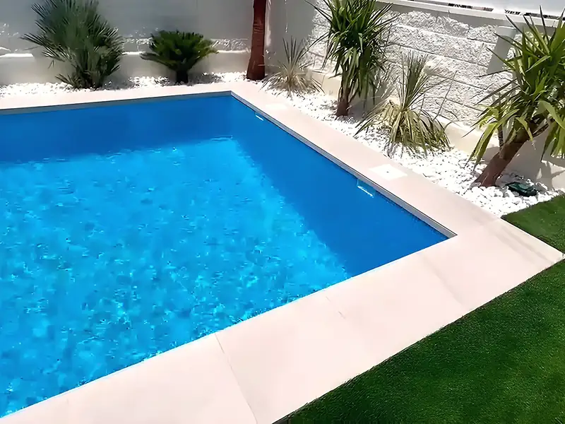 Rectangular pool with bright blue water, pale porcelain coping, white wall, young palms on white pebbles and artificial lawn