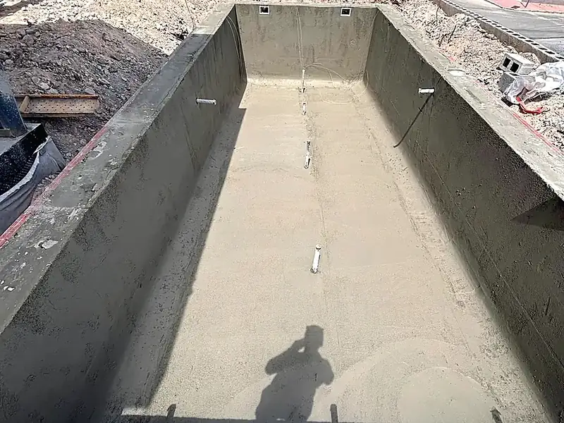 Concrete pool shell freshly rendered in grey cement, pipework for lights and inlets in place, excavated ground around it