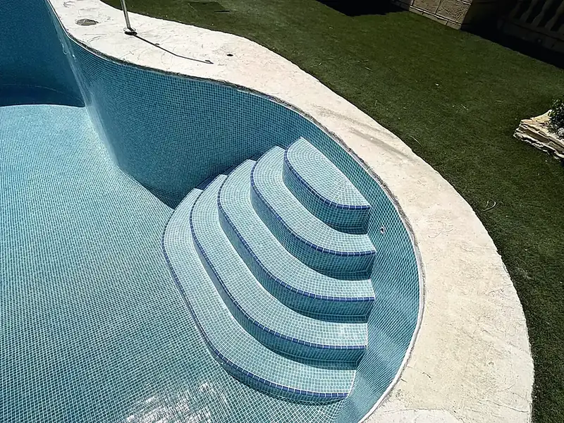 Empty freeform pool with pale blue mosaic, fan-shaped Roman steps, blue tile edging, cream stone coping and artificial lawn