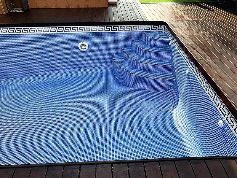 Empty rectangular pool with blue mosaic, Greek key waterline border, curved corner steps and dark timber decking all round