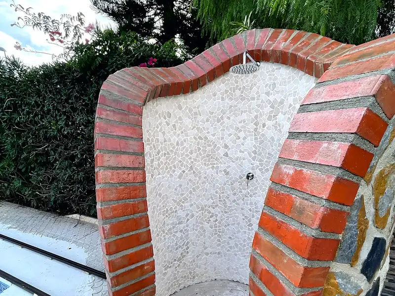 Outdoor shower niche in red brick with a curved white stone mosaic interior, rain head and natural stone cladding by a pool